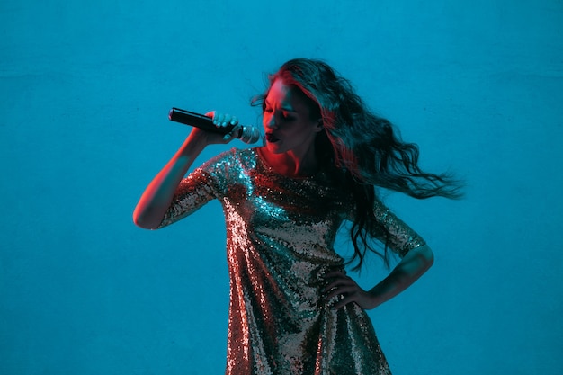 Retrato de cantante femenina caucásica aislado sobre fondo azul de estudio en luz de neón. Modelo de mujer hermosa en vestido brillante con micrófono. Concepto de emociones humanas, expresión facial, publicidad, música, arte.