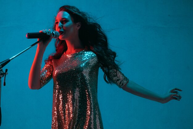 Retrato de cantante de raza caucásica aislado sobre fondo azul de estudio en luz de neón. Modelo de mujer hermosa en vestido brillante con micrófono. Concepto de emociones humanas, expresión facial, publicidad, música, arte.