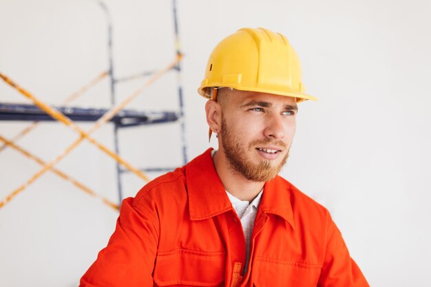 Retrato de capataz joven en ropa de trabajo naranja y casco amarillo con lápiz detrás de la oreja cuidadosamente mirando a un lado con andamios en el fondo