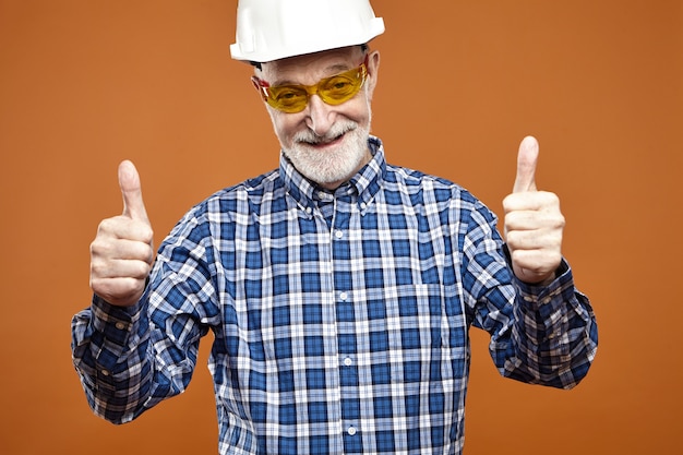 Retrato de capataz senior positivo guapo con casco y anteojos amarillos mostrando los pulgares hacia arriba gesto y sonriendo felizmente, animando a su equipo de trabajo para un buen trabajo. Construcción y renovación
