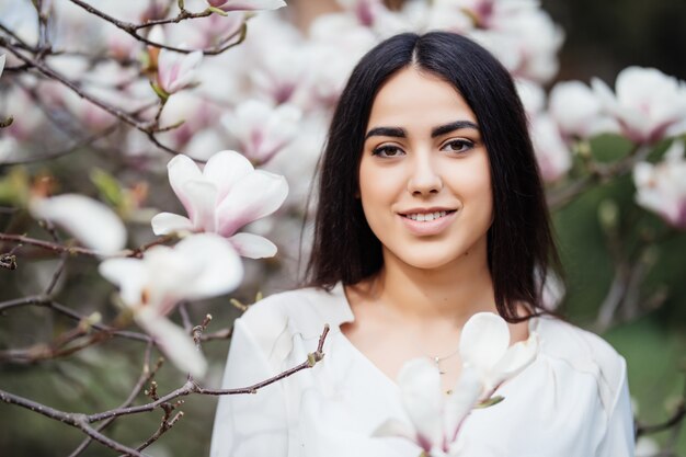 Retrato de cara de hermosa niña morena caucásica cerca del árbol de magnolia de flor al aire libre en el parque de la primavera.