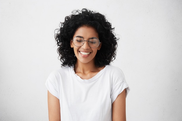 Retrato de carismática y encantadora joven africana con cabello rizado con gafas sylish, sonriendo ampliamente, entrecerrando los ojos