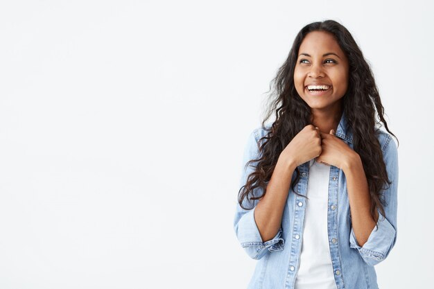 Retrato de una carismática y encantadora mujer afroamericana con el pelo largo y ondulado, vistiendo una elegante camisa de mezclilla, sonriendo ampliamente, emocionada de recibir la sorpresa de su novio, luciendo feliz.