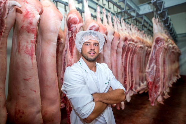 Retrato de carnicero profesional en la fábrica de almacenamiento en frío sosteniendo los brazos cruzados con la canal de cerdo en el fondo