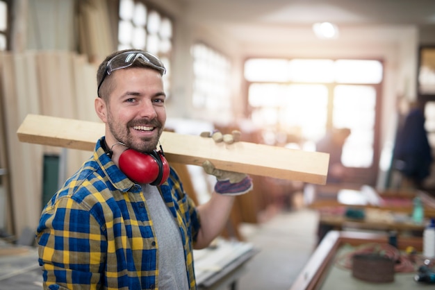 Retrato de carpintero profesional sosteniendo material de tablones de madera sobre su hombro y sonriendo