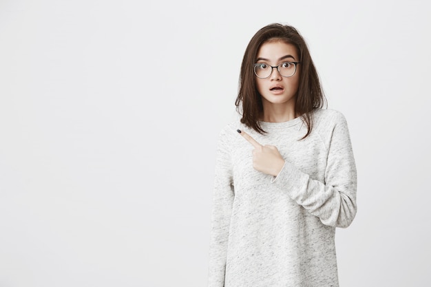 Retrato de caucásica linda morena con gafas, levantando las cejas con preocupación y conmoción, apuntando con el dedo índice a un lado