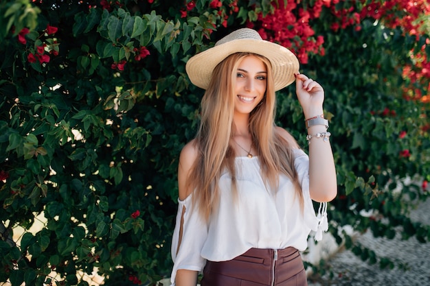 Retrato de cerca al aire libre de la joven y bella niña rizada sonriente feliz con elegante sombrero de paja en la calle cerca de rosas en flor Concepto de moda de verano. Copia espacio