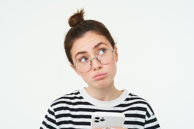Foto gratuita retrato de cerca de una chica con gafas mira desconcertada sostiene el teléfono y piensa hace una elección decide qué