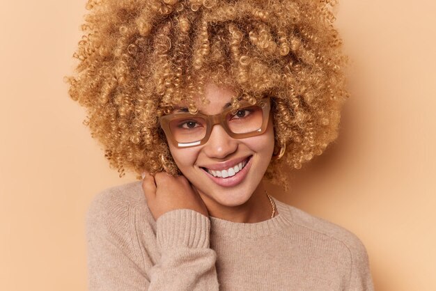 El retrato de cerca de una hermosa y alegre mujer joven de cabello rizado sonríe tiernamente mira directamente a la cámara y se siente satisfecho usa anteojos transparentes y un puente suave aislado sobre fondo marrón.