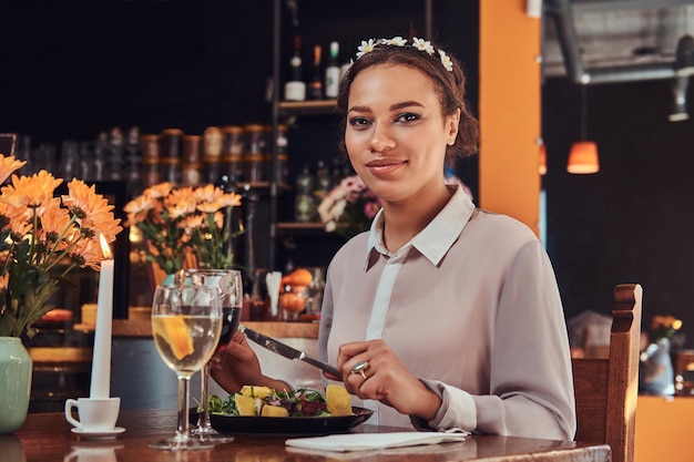 Retrato de cerca de una hermosa mujer de piel negra con una blusa y una diadema de flores, disfrutando de la cena mientras come en un restaurante.