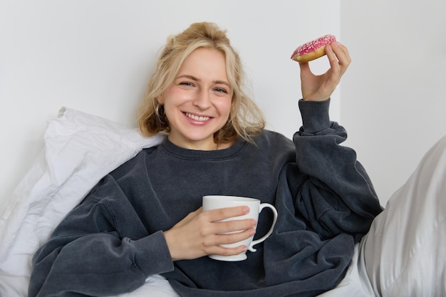 Foto gratuita retrato de cerca de una hermosa mujer rubia sonriente acostada en la cama bebiendo té y comiendo rosquillas en un