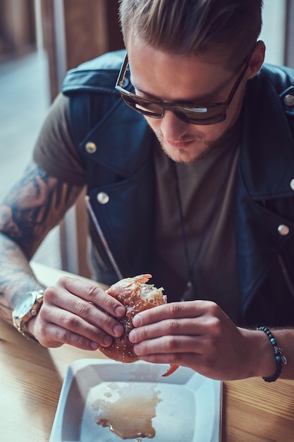 Retrato de cerca de un hipster hambriento con un elegante corte de pelo y barba sentado en una mesa, decidió cenar en un café al borde de la carretera, comiendo una hamburguesa.