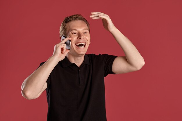 Retrato de cerca de un joven atlético jengibre con una elegante camiseta negra riéndose y hablando con un smartphone mientras posa en un estudio rosa. Expresiones faciales humanas. Emociones sinceras con