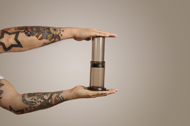 Retrato de cerca de las manos del hombre tatuado apretando un aeropress vacío en la pared gris claro