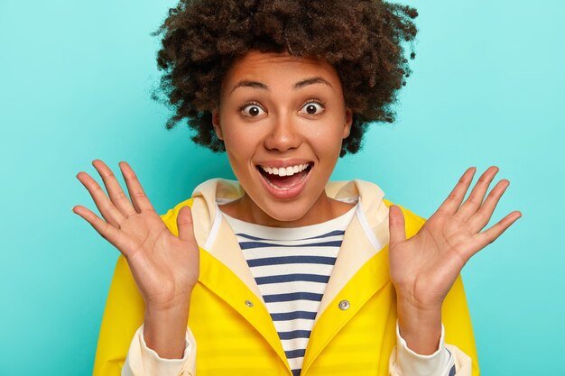 Retrato de cerca de una mujer alegre de piel oscura que levanta las palmas de las manos, se siente muy feliz, reacciona con sorpresa, sonríe ampliamente, muestra los dientes blancos, usa un impermeable amarillo y camina durante el tiempo de otoño aislado en azul