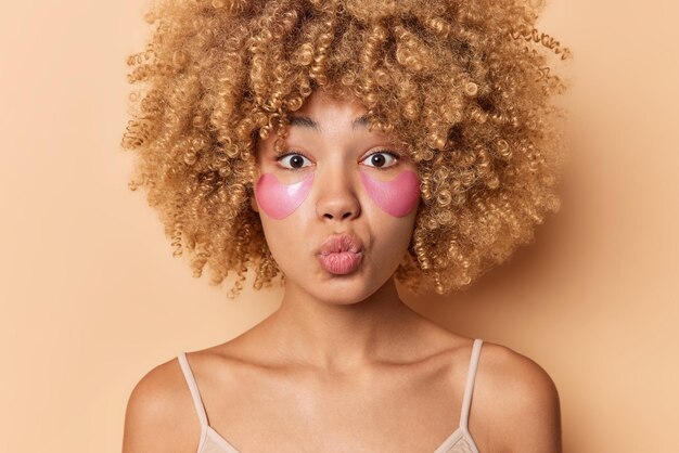 El retrato de cerca de una mujer de cabello rizado mantiene los labios doblados y quiere besar a alguien que aplica parches de hidrogel rosa debajo de los ojos para el rejuvenecimiento, usa una camiseta casual aislada sobre un fondo beige.