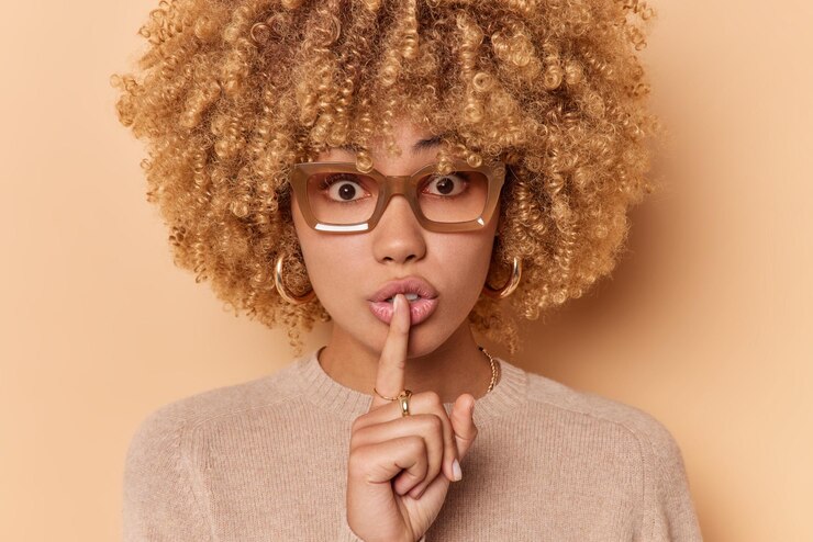 Foto gratuito el retrato de cerca de una mujer de pelo rizado hace un gesto de silencio que mantiene el dedo índice sobre los labios, tiene una expresión misteriosa, lleva gafas y un puente aislado sobre un fondo beige. lenguaje corporal
