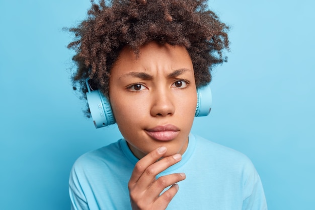 Retrato de cerca de una seria niña afroamericana mantiene la mano en la barbilla, parece disgustada vestida de manera informal, escucha música a través de auriculares o aprende nuevas palabras extranjeras aisladas sobre una pared azul