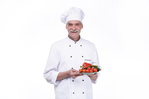 Retrato de chef sonriente sosteniendo un plato de verduras aislado en la pared blanca