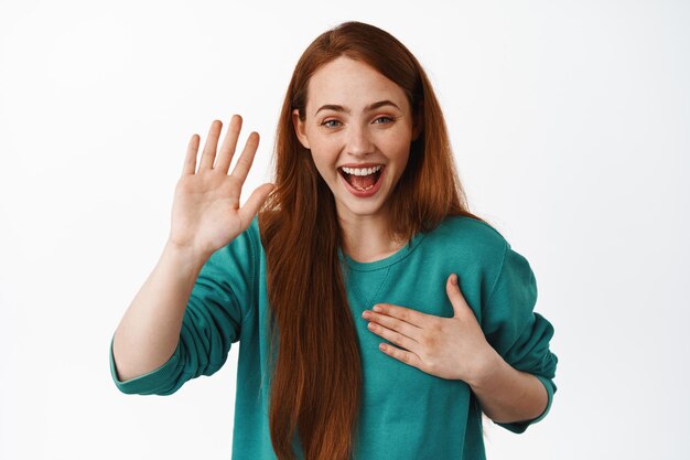 Retrato de una chica amigable y feliz, riendo y sonriendo, levantando la mano y colocando la palma en el corazón, hacer una promesa, ser honesta, presentarse, fondo blanco