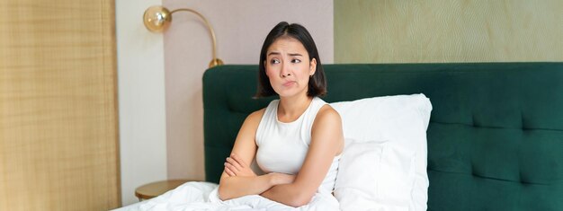 Foto gratuita retrato de una chica asiática en la cama con un aspecto complicado y preocupado, cruza los brazos en el pecho y frunce el ceño.