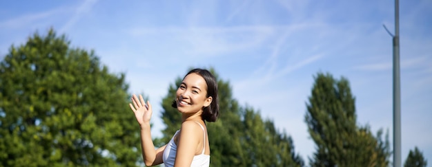 Foto gratuita retrato de una chica asiática en clase de fitness en el parque sentada en una alfombra de goma y agitando la mano a la cámara