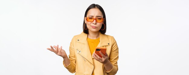 Retrato de una chica asiática confundida con gafas de sol sosteniendo un teléfono inteligente usando un teléfono móvil y luciendo desconcertada de pie sin idea sobre el fondo blanco