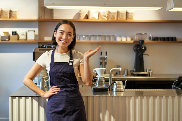 Retrato de una chica asiática en delantal parada en un café cerca del mostrador barista señalando con el dedo el espacio de la copia
