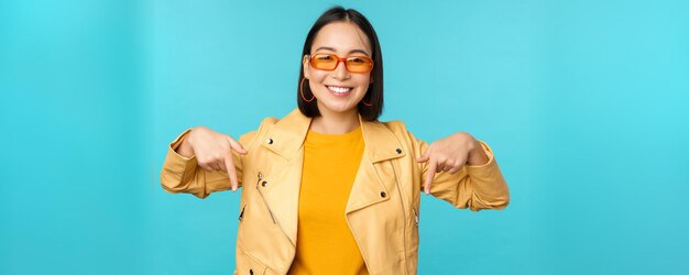 El retrato de una chica asiática con estilo usa gafas de sol, sonríe y señala con el dedo hacia abajo, muestra anuncios sobre fondo azul.