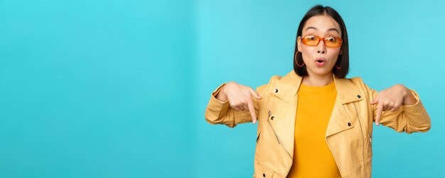 El retrato de una chica asiática con estilo usa gafas de sol, sonríe y señala con el dedo hacia abajo, muestra anuncios sobre fondo azul.