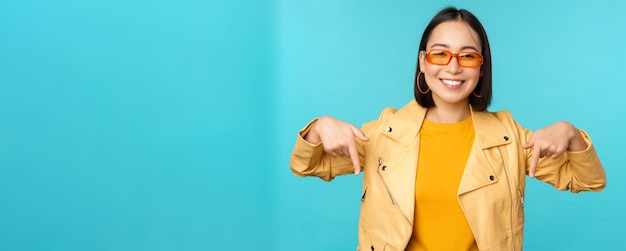 El retrato de una chica asiática con estilo usa gafas de sol, sonríe y señala con el dedo hacia abajo, muestra anuncios sobre fondo azul.