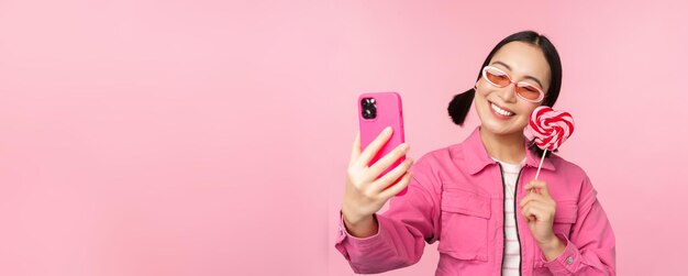 Retrato de una chica asiática feliz y elegante que se toma una selfie con dulces de lolipop y sonríe tomando una foto con una aplicación móvil de pie sobre un fondo rosa