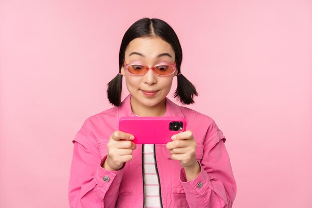 Retrato de una chica asiática feliz jugando en un teléfono inteligente viendo videos en una aplicación de teléfono móvil de pie sobre un fondo rosa