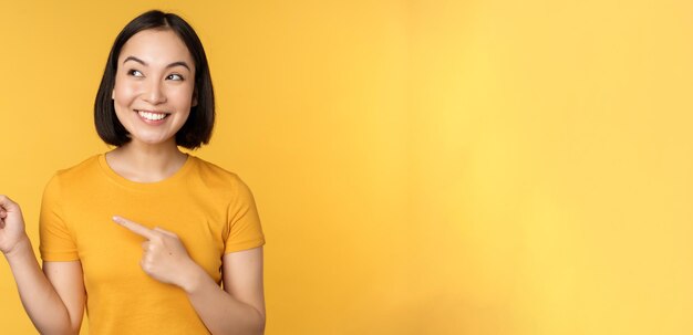 Retrato de una chica asiática feliz señalando con el dedo y mirando a la izquierda sonriendo asombrada mirando el cartel promocional que muestra publicidad contra el fondo amarillo