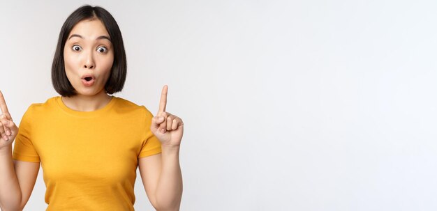 Retrato de una chica asiática impresionada que señala con el dedo hacia arriba dice wow mirando asombrada a la cámara de pie sobre fondo blanco Copiar espacio