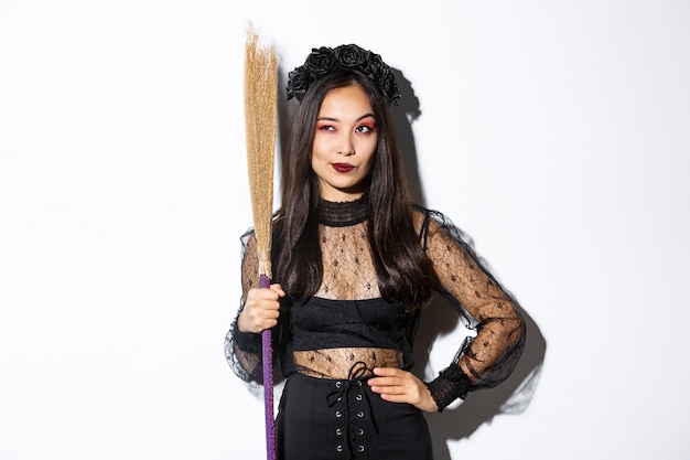 Retrato de una chica asiática inteligente y reflexiva mirando la esquina superior izquierda con una sonrisa satisfecha, sosteniendo una escoba, vistiendo un disfraz de bruja para la fiesta de Halloween, de pie sobre un fondo blanco