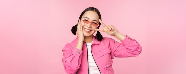 Retrato de una chica asiática kawaii con gafas de sol que muestra un signo de paz cerca de los ojos y sonríe lindo a la cámara p