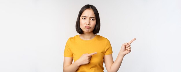 Retrato de una chica asiática malhumorada decepcionada señalando con el dedo a la derecha y mirando algo injusto quejándose de pie sobre fondo blanco