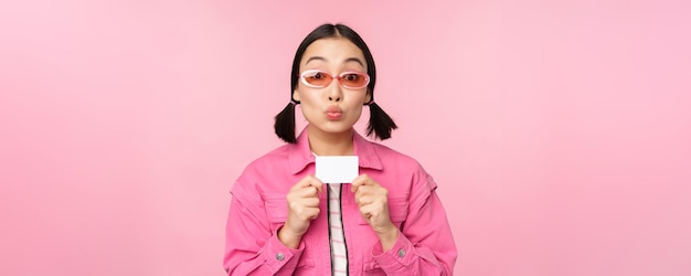 El retrato de una chica asiática moderna y elegante muestra una tarjeta de crédito con descuento y se ve complacida pagando el concepto sin contacto de comprar de pie sobre un espacio de copia de fondo rosa