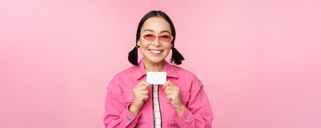 El retrato de una chica asiática moderna y elegante muestra una tarjeta de crédito con descuento y se ve complacida pagando el concepto sin contacto de comprar de pie sobre un espacio de copia de fondo rosa