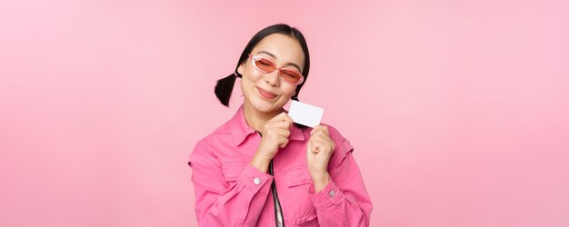 El retrato de una chica asiática moderna y elegante muestra una tarjeta de crédito con descuento y se ve complacida pagando el concepto sin contacto de comprar de pie sobre un fondo rosa