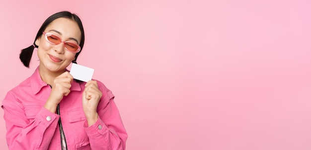 Foto gratuita el retrato de una chica asiática moderna y elegante muestra una tarjeta de crédito con descuento y se ve complacida pagando el concepto sin contacto de comprar de pie sobre un fondo rosa