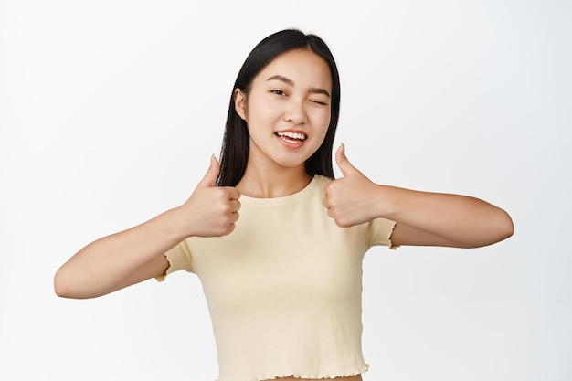 Retrato de una chica asiática morena sonriente mostrando los pulgares hacia arriba guiñando un ojo y mirando a la cámara feliz animándote a pararte sobre fondo blanco