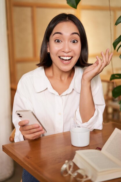 Foto gratuita retrato de una chica asiática que parece sorprendida sosteniendo un teléfono móvil con cara de asombro buenas noticias en smartph