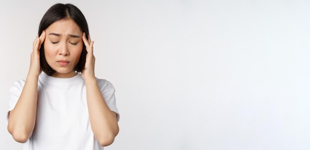 Retrato de una chica asiática que siente dolor de cabeza, migraña o está enferma de pie con una camiseta blanca sobre una ba blanca