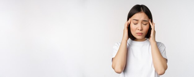 Retrato de una chica asiática que siente dolor de cabeza, migraña o está enferma de pie con una camiseta blanca sobre una ba blanca