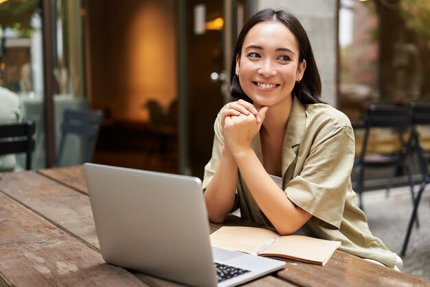 Foto gratuita retrato de una chica asiática que trabaja al aire libre en un café se sienta con estudios portátiles sonríe feliz