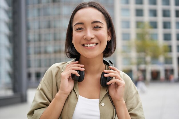 Retrato de una chica asiática sonriente con auriculares posando en el centro de la ciudad escuchando música