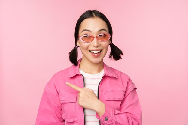 Retrato de una chica asiática sonriente con elegantes gafas de sol apuntando con el dedo a la izquierda mostrando una pancarta publicitaria de pie sobre fondo rosa Copiar espacio