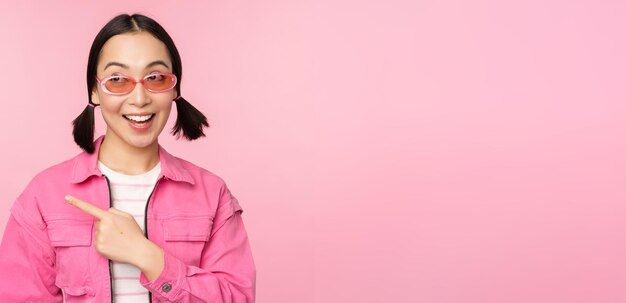 Retrato de una chica asiática sonriente con elegantes gafas de sol apuntando con el dedo a la izquierda mostrando una pancarta publicitaria de pie sobre fondo rosa Copiar espacio